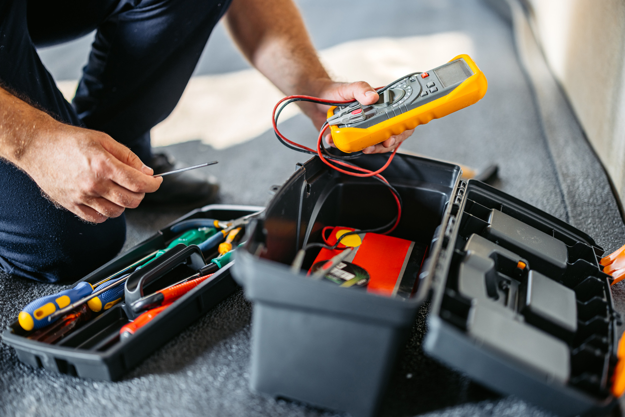 Person using a multimeter from a toolbox filled with tools on a work surface.
